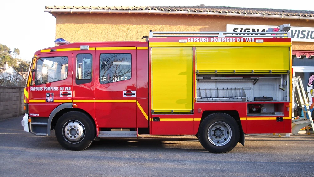 Gros plan ou vue trois-quarts d'un véhicule utilitaire blanc avec marquage professionnel en graphisme coloré appliqué sur le flanc, montrant des éléments visuels nets (logo, texte, design graphique), photographié en lumière naturelle de jour sur un parking ou en milieu urbain.